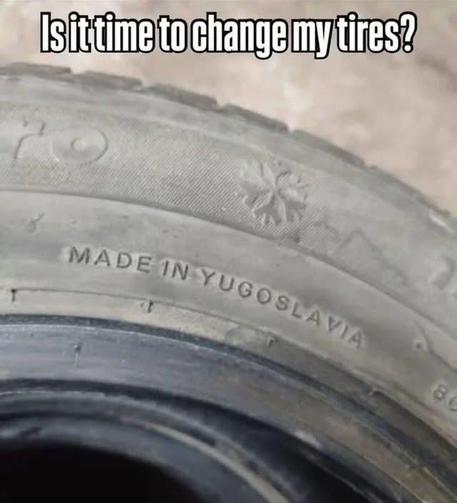 The image is a close-up, eye-level shot of a worn tire. Text is printed on the sidewall of the tire. At the top of the image, a white text overlay asks, "Is it time to change my tires?". The tire itself is predominantly dark gray and appears to be a winter tire due to a small, embossed snowflake symbol visible on its surface. Below the snowflake, the words "MADE IN YUGOSLAVIA" are clearly legible, also embossed into the rubber. The texture of the tire suggests age and wear, with the tread pattern not fully visible due to the close cropping. The background is out of focus and appears to be a light, neutral color, possibly concrete or dirt, providing no significant distraction from the tire. The composition focuses heavily on the text and the snowflake symbol, highlighting the origin of the tire and posing a question about its condition.